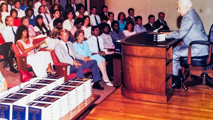 Herbert W. Armstrong at a desk before rows of students, stacks of books in front of him