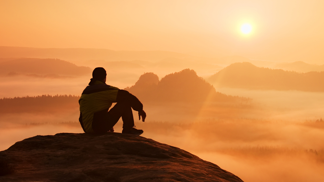 Hiker in black on the rocky peak. Wonderful daybreak in mountains, heavy orange mist in deep valley. Man sit on the rock and watch over the fog .