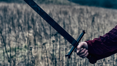 Man with bead and long hair stands outside. He wears historic clothes and could be a vking or a knigth. He holds a metal helmet under one of his arms. In his right hand he holds a sword.