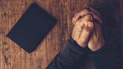 Christian man praying with hands crossed and Holy Bible by his side on wooden desk in church, top view