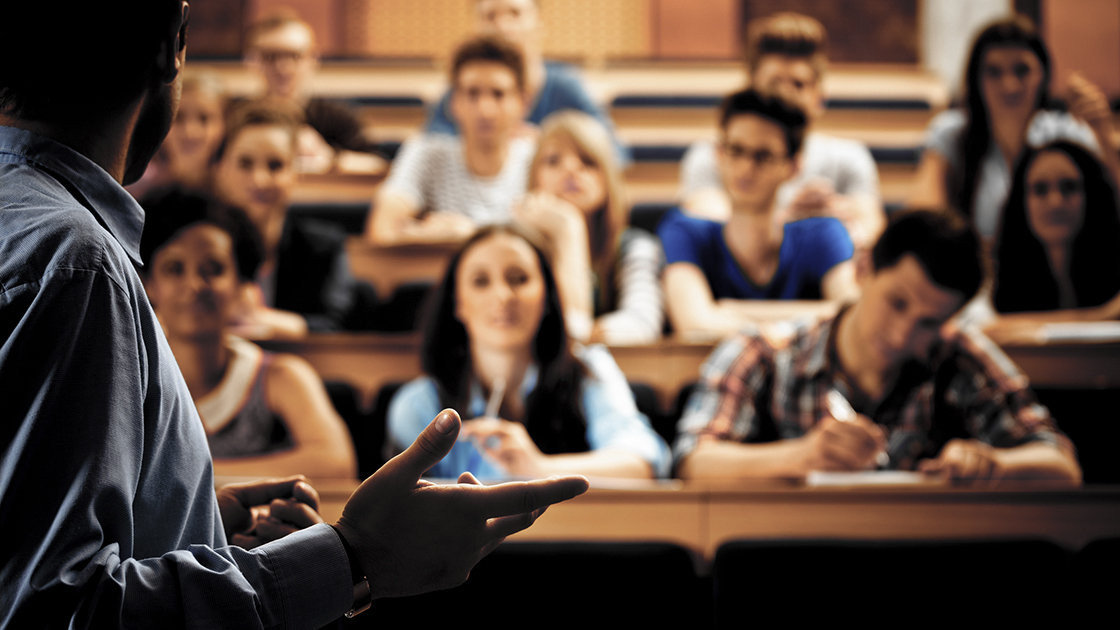 Large group of students sitting in the lecture hall at university and listening to their teacher. Focus on the professor. Dark tones.