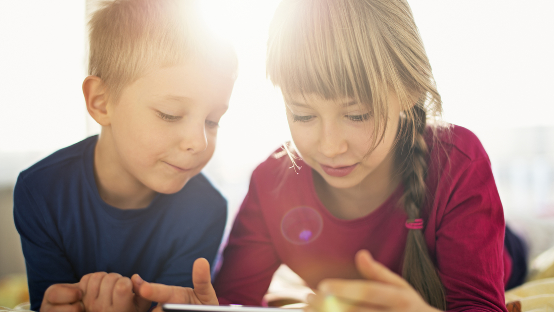 Brother and sister using digital tablet. Kids are aged 4 and 7 and are lying on a bed on a sunny day.