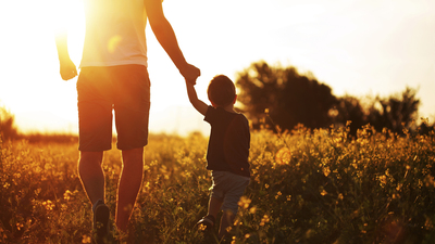 Photo of little boy holding his father hand and walking at the field in the sunset