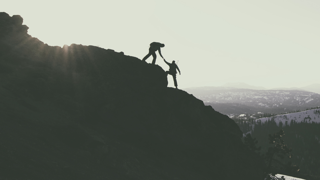 Two strong hikers, one helping the other up a ridge to the summit as the sun sets behind them. 