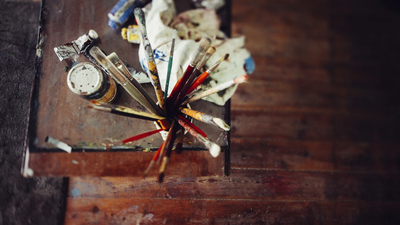 Topview of a wooden table in an art studio with a collection of paintbrushes in a container and tins and tubes of paint alongside a rag