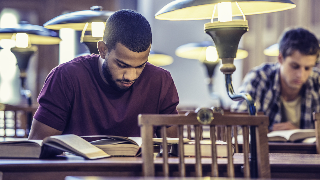 Two male students sitting behind old-fashioned desks with lamps, reading from their books.