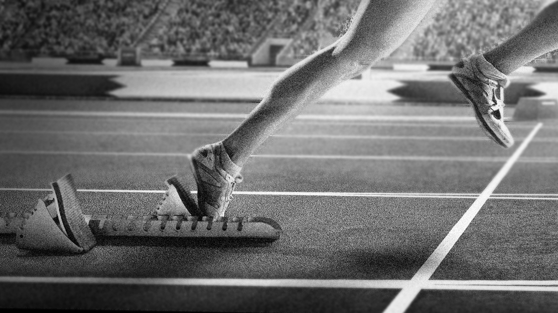 Professional female athlete sprinting from blocks on numbered start line on outdoor athletics track on olympic stadium full of spectators under a dramatic evening sky. Sprinter is wearing generic athletics kit.