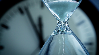 A hourglass with falling sand in front of a clock reaching midnight. Concept photo urgency, and time is running out and deadline is approaching. Close-up of hour glass is photographed in horizontal format with copy space, against a soft-focus clock face in the background.