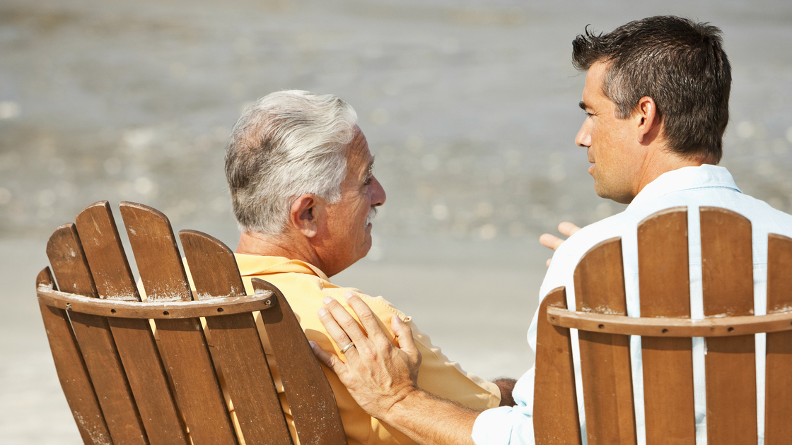 Rear view of senior man (60s) talking with adult son (40s), sitting on adirondack chairs on the beach.