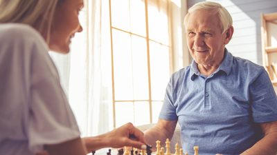 Beautiful young girl-volunteer and handsome old man are playing chess and smiling