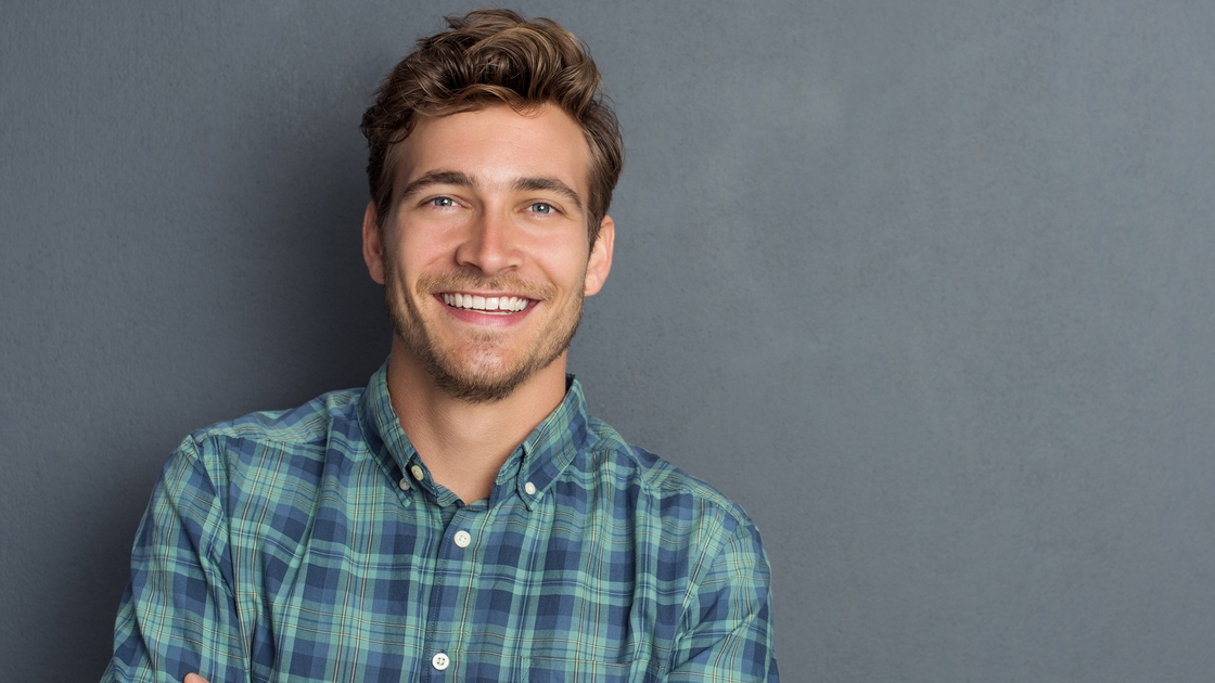 Young handsome man leaning against grey wall with arms crossed. Cheerful man laughing and looking at camera with a big grin. Portrait of a happy young man standing with crossed arms over grey background.