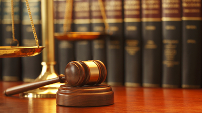 A gavel rests on sounding block next to a brass scale of justice in front of a long row of law books on a desk in a law office. Photographed with a very shallow depth of field.