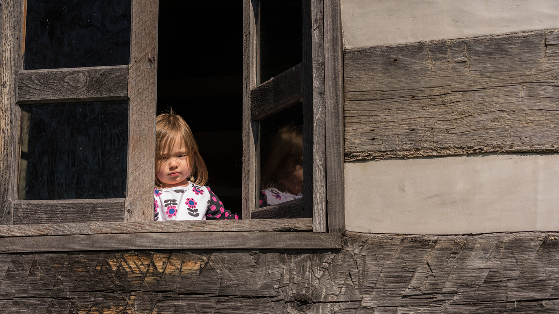 Small and lonely caucasian baby girl or toddler looking out of old wooden cabin window to suggest poverty or recession