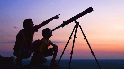 A young boy looking thru a telescope at sunset with his father.