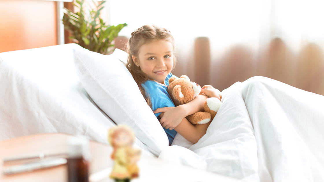 Cute little girl lying in hospital bed with teddy bear and smiling at camera