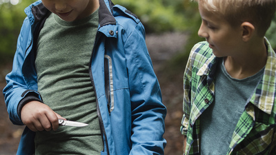 Young Boy showing off and pulling a knife out from underneath his jacket white his friends stand around in a woodland area.