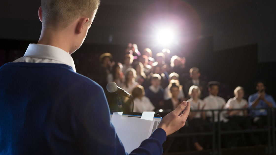 Male student making a speech. He is bstanding at the podium and is talking to the crowd of people.