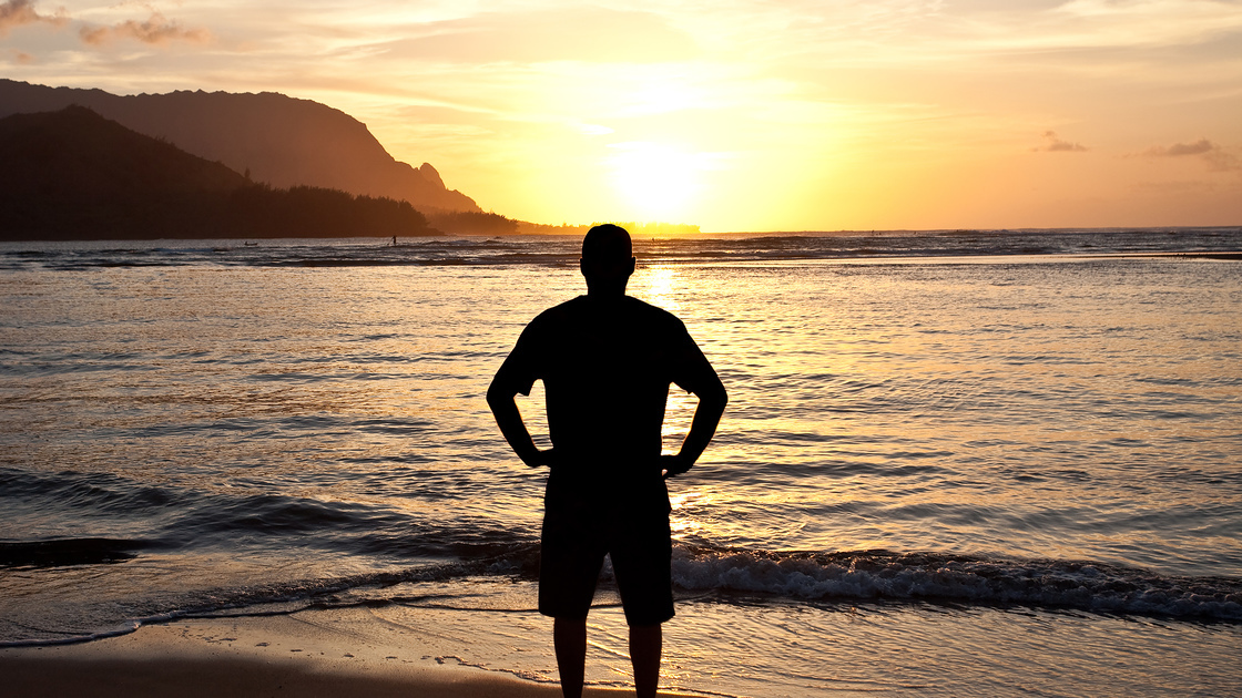 A silhouette of a man watching a beautiful sunset over the ocean. Themes include watching the sunset, nature, beauty, men, single, alone, contemplation, Hawaii, beach, resort, tropical, and yoga. Man is unrecognizable and in his mid 30s. 