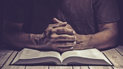 Man praying on a wooden table with an open Bible.