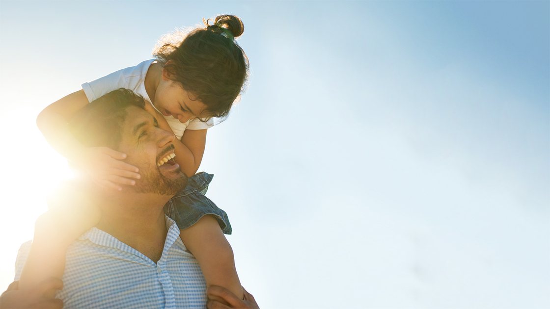 A young girl sits on her dad's shoulders, looking at each other, both smiling outdoors in the sun