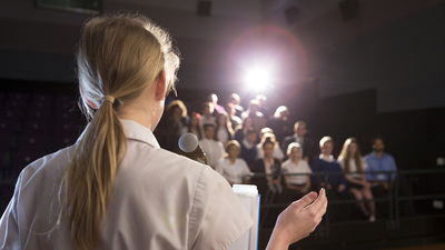 Female student making a speech. She is standing at a podium and smiling to the crowd.