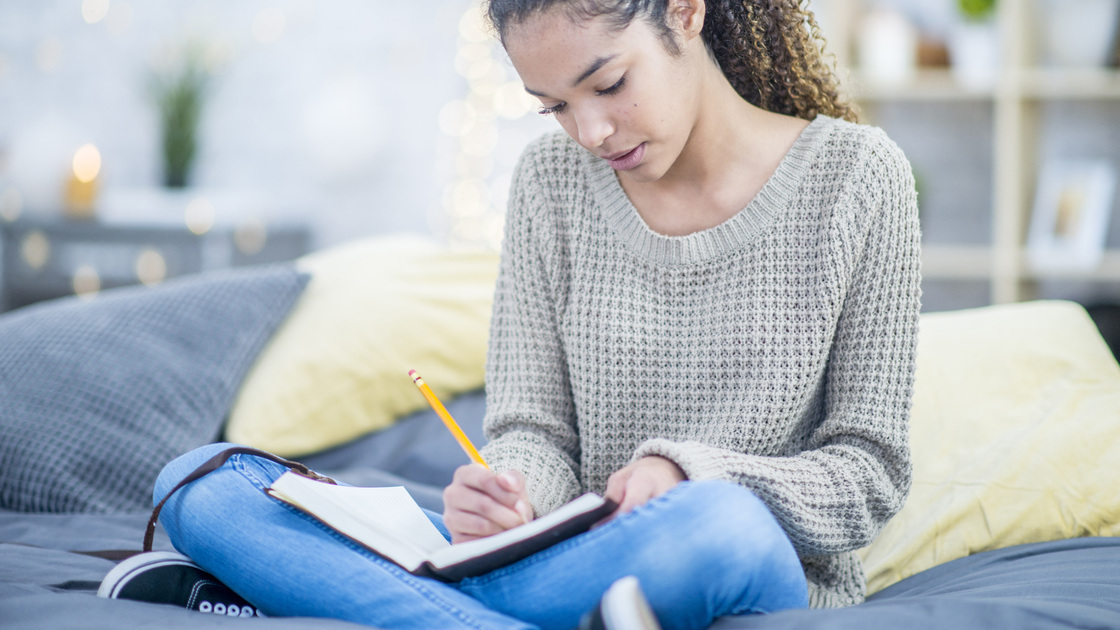 A pretty teenaged girl writes into a journal with interest while sitting cross legged on her bed.