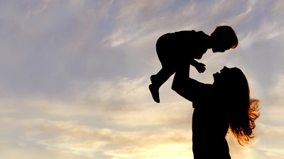 A silhouette of a happy young mother, laughing as she plays with her toddler child and lifts him over her head outside, isolated against the sunset.