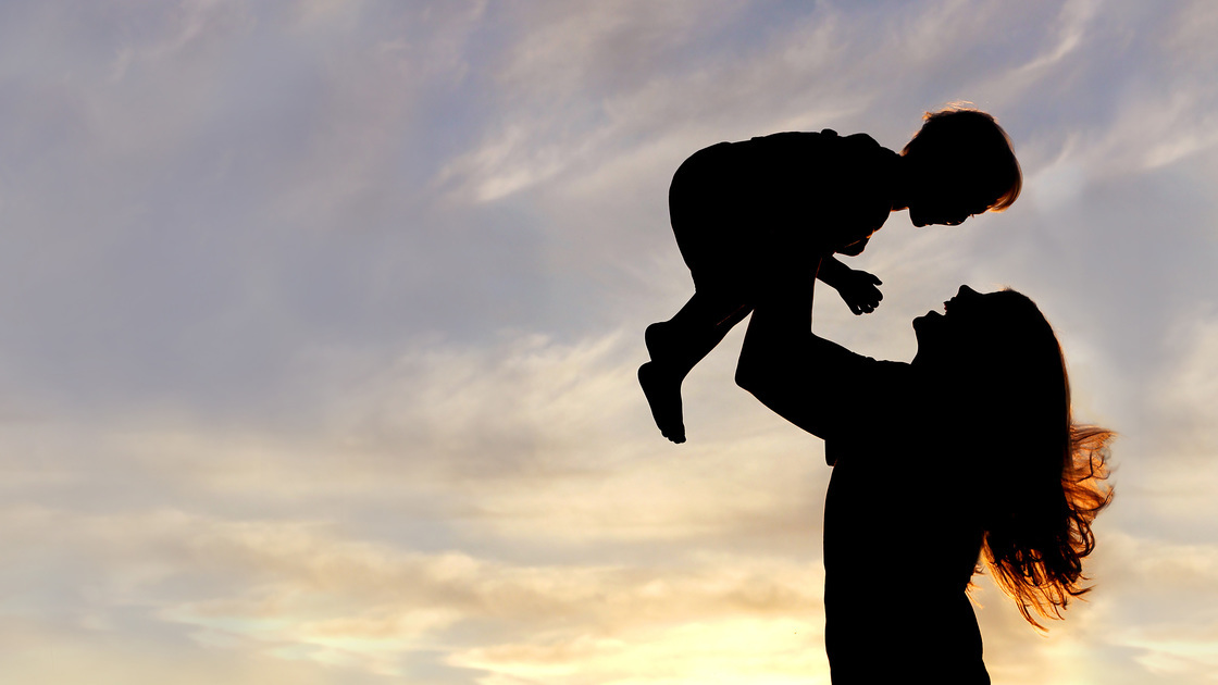 A silhouette of a happy young mother, laughing as she plays with her toddler child and lifts him over her head outside, isolated against the sunset.