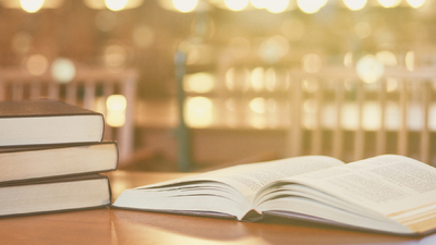 Open book on a desk in library's reading room. Electric lamps and stack of books in the frame.