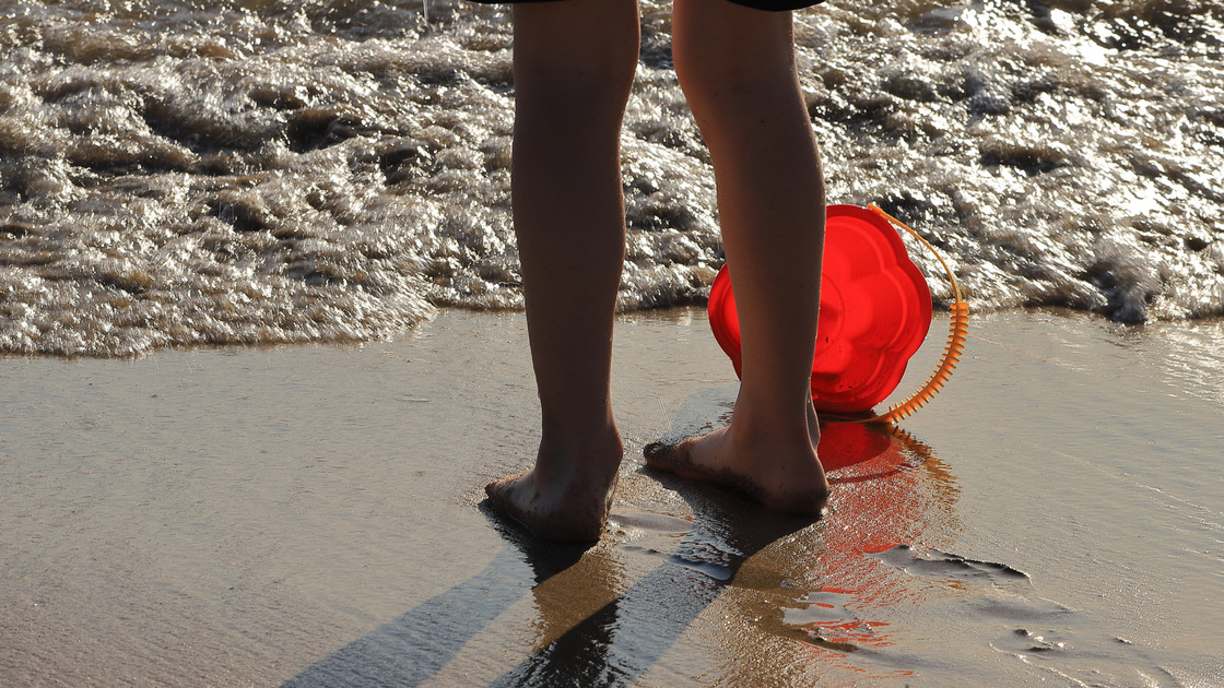 A child children's feet on the beach plays with water with a toy bucket