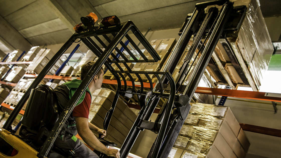 Wide shot of warehouse worker lifting a pallet of goods with a forklift in a colorful warehouse setting. Low angle view.