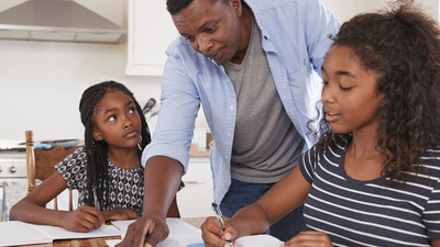 Father Helping Two Daughters Sitting At Table Doing Homework