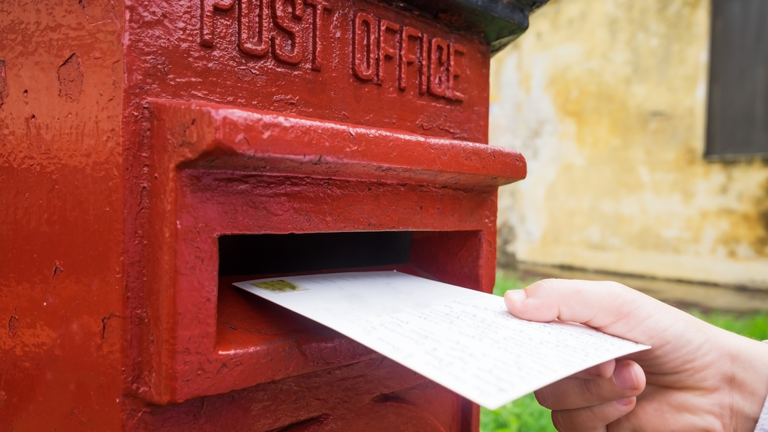 Closeup on a male hand putting a letter in a red letterbox. Concept of vintage type of communication. To send postcard from vacation or travel