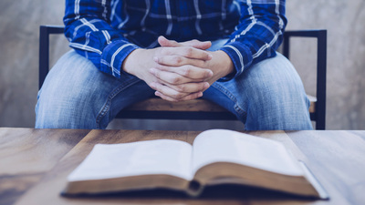 A man sitting on wooden chair praying to God with blurred open bible on wooden table foreground, trust concept