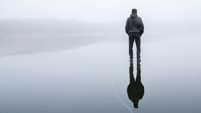Young adult man standing alone on cracked dark ice surface. Mist over frozen lake in winter. Foggy air. Early chilly morning. Peaceful atmosphere in nature. Back view.