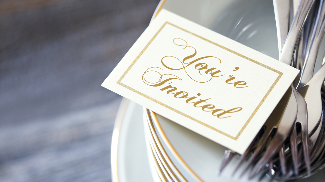 An invitation and a grouping of silverware rest on top of a stack of dinner plates. The image is photographed using a very shallow depth of field.