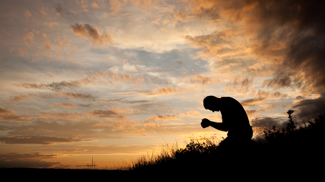 A silhouette of a man praying in a meadow. Side view of Caucasian man folding his hands and meditating. Themes include spirituality, god, religion, asking, pleading, heaven, faith, salvation, forgiveness, and discipleship. Man, in his 40s, is unrecognizable in the image. 