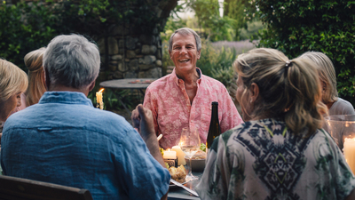 A group of mature friends are sitting around an outdoor dining table, eating and drinking. They are all talking happily and enjoying each others company. The image has been taken in Tuscany, Italy.