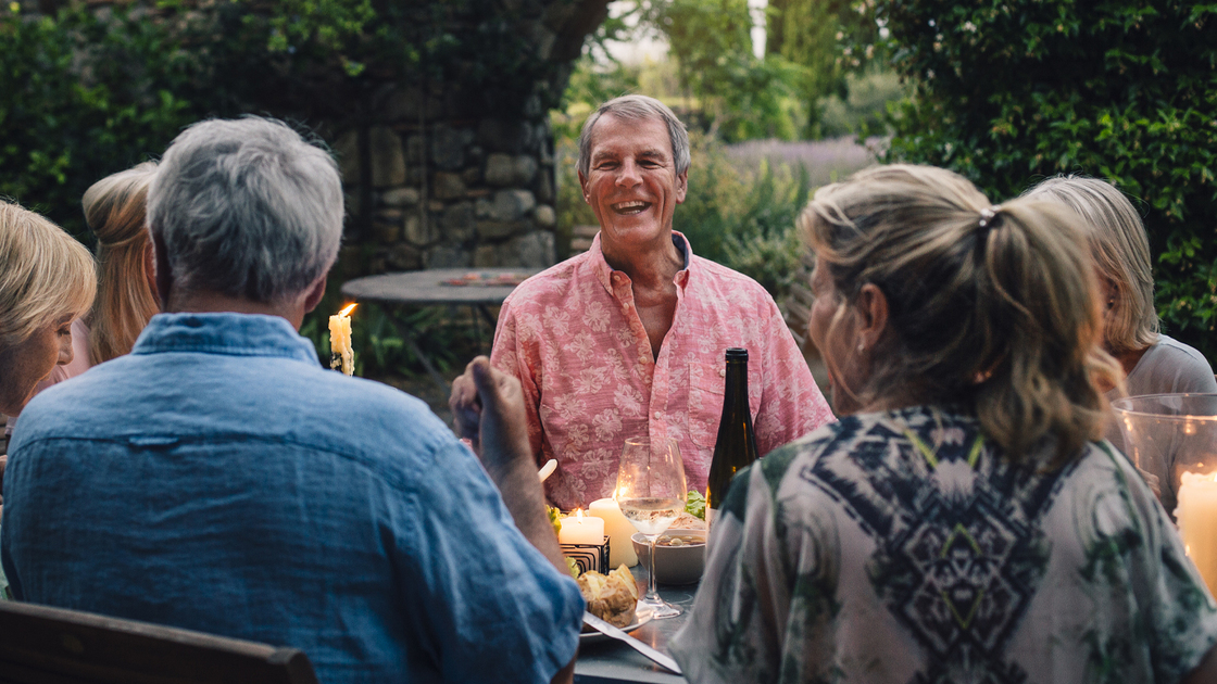 A group of mature friends are sitting around an outdoor dining table, eating and drinking. They are all talking happily and enjoying each others company. The image has been taken in Tuscany, Italy.