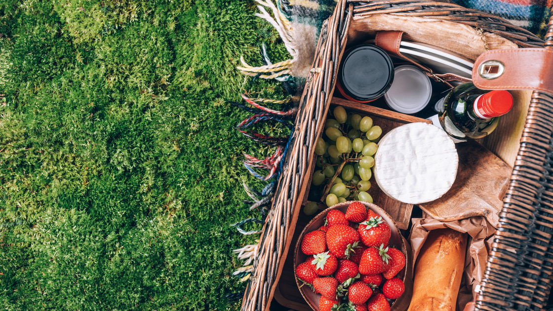 Picnic set with fruit, cheese, honey, strawberries, grapes, baguette, wine, wicker basket for picnic on plaid over green grass. Top view. Copy space. Summer picnic time, family lunch. Romantic picnic.