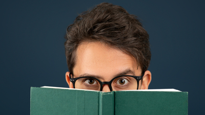 Young man wearing eyeglasses peeking over the top of green book, nerdy male showing curiosity and interest, standing isolated on dark blue studio background, enjoying reading, closeup