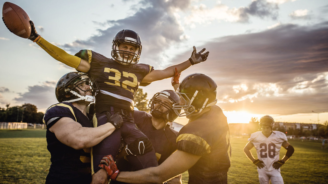 American football players holding their teammate high up on a playing field while celebrating a victory.