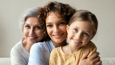 Caring grown woman mother hold on knees hug girl kid while mature female grandmother pensioner embrace her adult daughter from back. Family portrait of women dynasty look at camera sit on sofa cuddle
