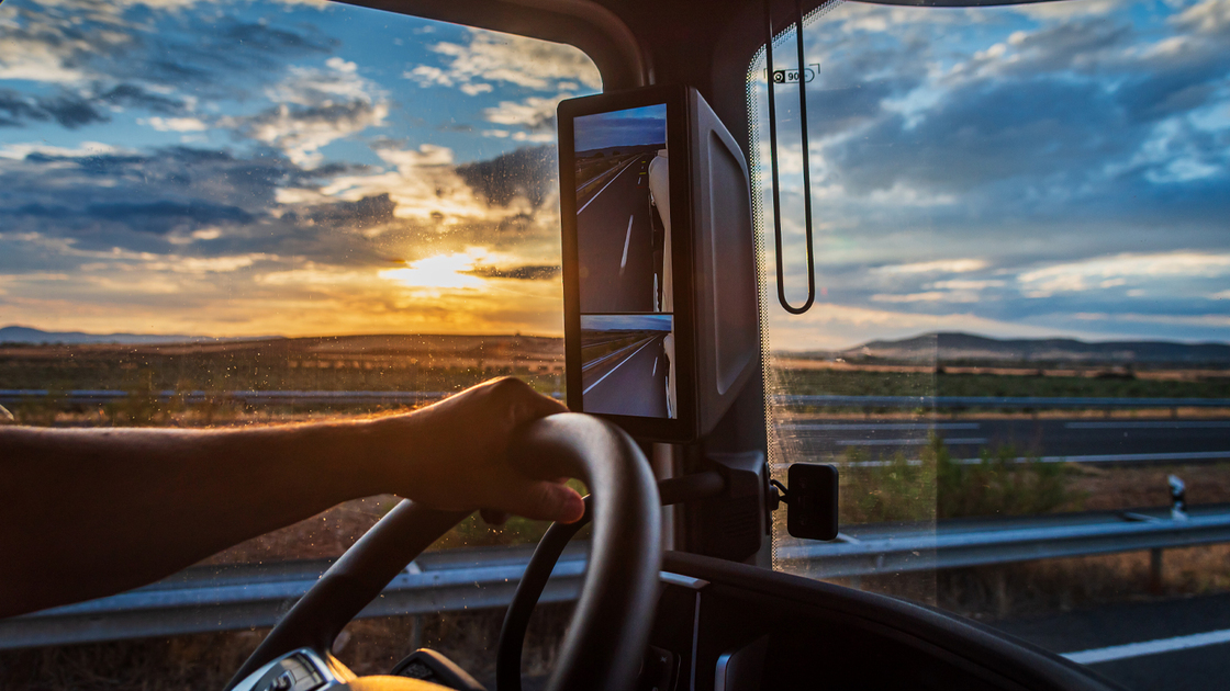 Truck cabin view with driver's hand on the wheel under a colorful sunset sky